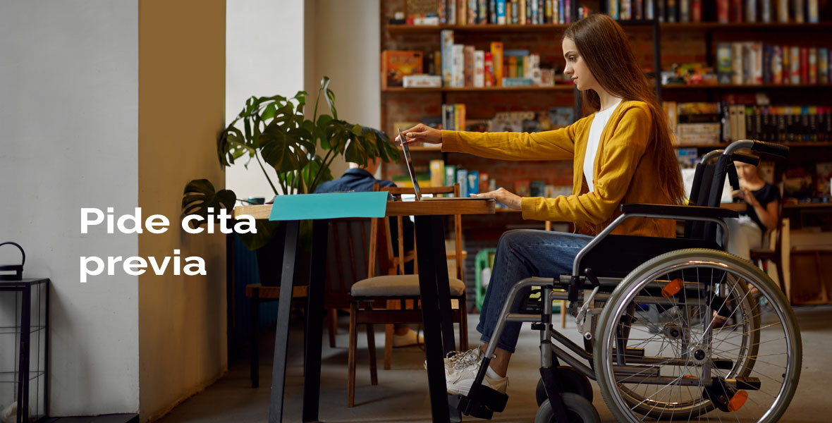 Mujer en silla de ruedas mirando un ordenador portátil en una mesa y al fondo estantería con libros. Hay un rotulo de 